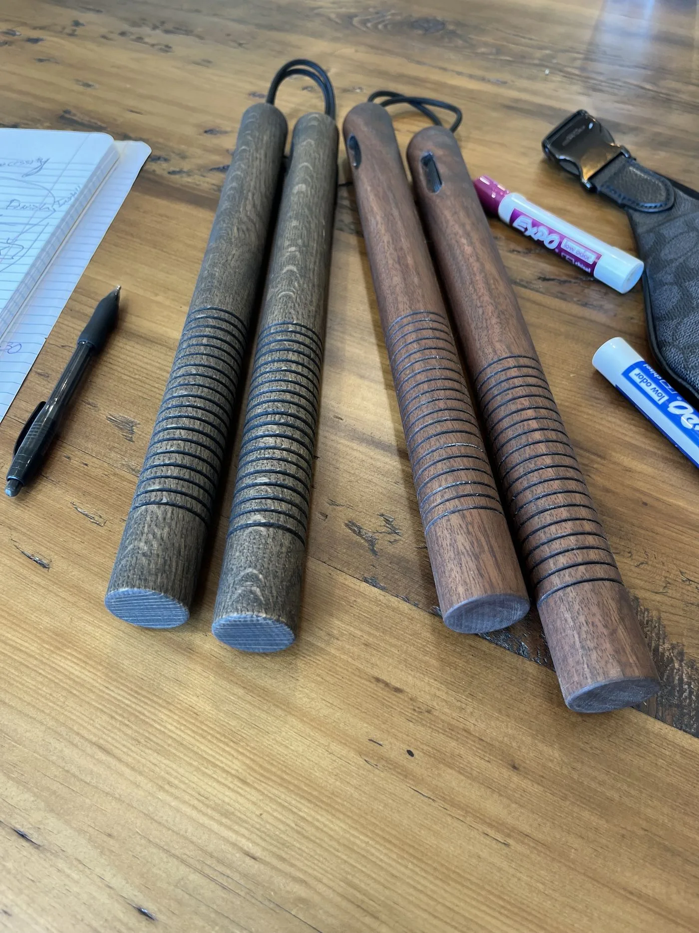 Two finished handmade nunchaku pairs on a reclaimed wood table — white oak with a black stain on the left, walnut on the right